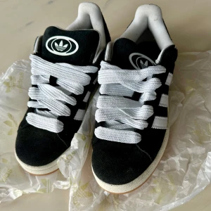 Svarta Adidas Campus sneakers - Snygga svarta Adidas Campus sneakers med vita detaljer och klassiska tre ränder på sidan. Skorna har en bekväm passform och är tillverkade i mocka med vita snören och en vit sula. Perfekta för en stilren look.