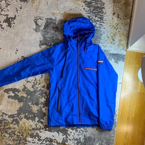 Vindjacka  - Säljer en snygg blå vindjacka.         Jackan har en sällsynt marinblå färg samt huva och dragkedja framtill.  Passar perfekt för vår och höst.           Den är i ny skick och har inte använts. Skriv för mer info!