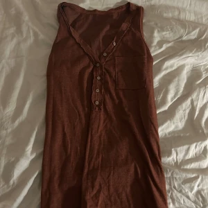 Brun/vinröd ärmlös linnetopp - Säljer en brun ärmlös linnetopp med knappar framtill och en bröstficka. Perfekt för varma dagar och enkel att matcha med andra plagg.