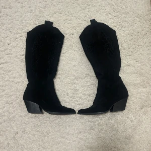 Svarta cowboy boots i mocka - Snygga svarta cowboy boots i mocka med dragkedja på sidan och en stabil klack. Dessa har kommit till användning några gånger vilket gör att klacken på nära håll fått lite slitage. De är annars i väldigt bra skick och passar till allt.