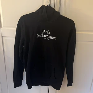 Svart hoodie från Peak Performance - Säljer en stilren svart hoodie från Peak Performance med vit logga på bröstet. Tröjan har långa ärmar och en klassisk huva. Perfekt för en avslappnad look.
