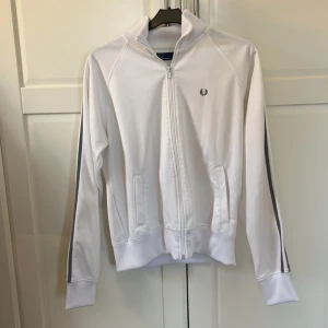 Vit track jacket från Fred Perry - Säljer en stilren vit track jacket från Fred Perry med grå ränder längs ärmarna. Jackan har dragkedja framtill och två fickor. Perfekt för en sportig look eller avslappnad stil.