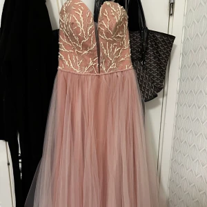 Rosa långklänning med broderad överdel - Elegant rosa långklänning med en vacker broderad överdel i guld. Klänningen har en axelbandslös design och en luftig tyllkjol som är öppen vid sidan. Perfekt nu till balen, klänningen är i väldigt bra skick då den användes bara en gång. hör av dig om du är intresserad, 