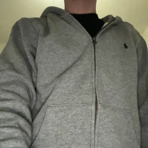 Grå hoodie från Ralph Lauren - Säljer en klassisk grå hoodie från Ralph Lauren med dragkedja och en liten logotyp på bröstet. Perfekt för en avslappnad stil. Den har långa ärmar och en bekväm huva.