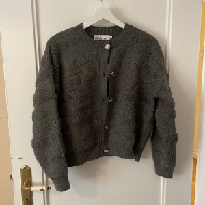 Grå kofta från Karen by Simonsen - Säljer en snygg grå kofta från Karen by Simonsen. Koftan har ett unikt texturerat mönster och knäpps med stiliga knappar framtill. Perfekt för att hålla sig varm och trendig under kyligare dagar.