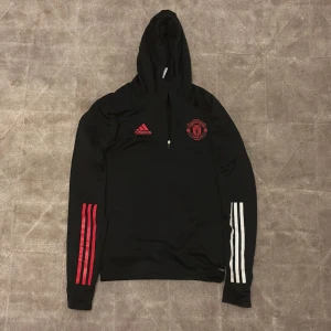 Svart hoodie från Adidas med Manchester United-logga - Säljer en svart hoodie från Adidas med Manchester United-logga på bröstet. Tröjan har en dragkedja vid halsen och långa ärmar med röda och vita ränder. Perfekt för fotbollsfans! Kom med prisförslag!