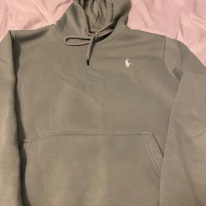  hoodie från Ralph Lauren - Säljer en stilren grön hoodie från Ralph Lauren med klassisk logga på bröstet. Den har en bekväm passform och en praktisk känguruficka framtill. Perfekt för en avslappnad stil. Hör av dig vid intresse!