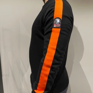 Svart tröja med orange detaljer från Parajumpers - Snygg svart tröja från Parajumpers med orange ränder längs ärmarna. Tröjan har en rund halsringning och ett märke på ärmen. Storlek M. Tar inte emot returer. 