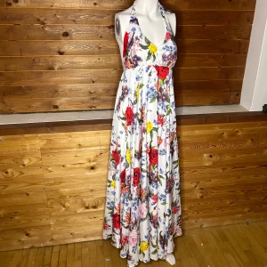 Blommig långklänning med knytband - Säljer en vacker långklänning med blommigt mönster i rött, blått och gult på vit botten. Klänningen har en v-ringad hals och knytband vid axlarna för en justerbar passform. Perfekt för sommarens alla tillfällen! Kan knytas på två olika sätt som på bilden och halterneck 