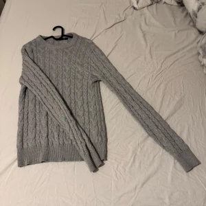 Grå stickad tröja - Säljer en stilren grå stickad tröja med kabelstickat mönster. Tröjan har långa ärmar och ribbade muddar vid ärmslut och nederkant. Perfekt för kyligare dagar! Pris kan diskuteras!