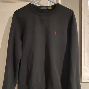  Ralph Lauren tröja - Säljer en stilren svart tröja från Ralph Lauren med det klassiska röda logotypen på bröstet. Tröjan har långa ärmar och en rund halsringning. Perfekt för en avslappnad och snygg look.