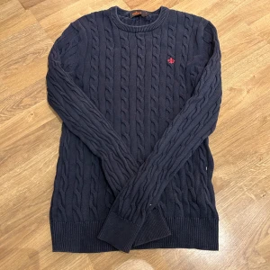 Morris tröja - Säljer en snygg mörkblå stickad tröja från Morris med kabelstickat mönster. Tröjan har en liten röd broderad detalj på bröstet och ribbade muddar vid ärmslut och nederkant. Perfekt för kyliga dagar!