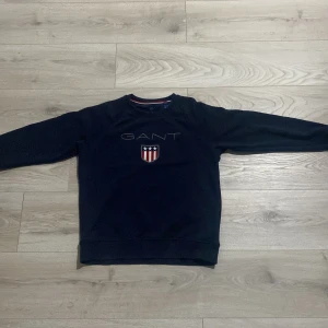 Mörkblå tröja från GANT - Säljer en snygg mörkblå tröja från GANT med broderad logotyp och ett stilrent emblem på bröstet. Tröjan har långa ärmar och en klassisk rund halsringning med röd och vit randig detalj på insidan av kragen.