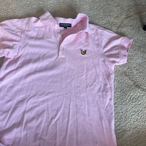 Rosa pikétröja från Lyle & Scott - Snygg rosa pikétröja från Lyle & Scott med klassisk krage och knappslå. Tröjan har en broderad logotyp på bröstet och korta ärmar. Perfekt för en avslappnad stil. STORLEK S
