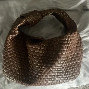 Brun flätad axelväska - Snygg brun axelväska med flätat mönster. Väskan har en bred axelrem som gör den bekväm att bära. Passar utmärkt för både vardag och speciella tillfällen.hyfsat rymlig.köpte väskan från plick 