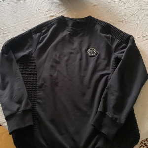 Svart tröja från Philipp Plein - Snygg svart tröja från Philipp Plein med unika quiltade detaljer på sidorna och en dragkedja. Tillverkad i en blandning av bomull och polyester för en bekväm passform. Perfekt för en stilren look. Använd få gånger då tröjan inte var i min storlek