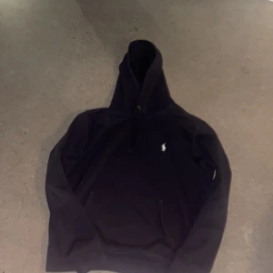 Svart hoodie från Ralph Lauren - Säljer en stilren svart hoodie från Ralph Lauren med det klassiska logotypbroderiet på bröstet. Tröjan har en bekväm passform och en praktisk huva med dragsko. Perfekt för en avslappnad look.