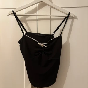 Svart linnetopp från Gina Tricot - Snygg svart linnetopp från Gina Tricot med smala axelband och en söt rosett framtill. Toppen har en liten rynkdetalj vid bysten och spetskant längs urringningen. Perfekt för en stilren look. I satin