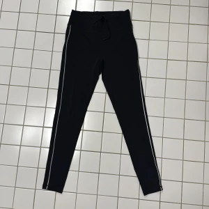 Svarta leggings från RS Sports - Snygga svarta leggings från RS Sports med vita/reflex  längs sidorna. Dold ficka i midjebandets mitt bak för mobil eller nycklar. Tillverkade i en mix av polyamid och lycra för en bekväm och stretchig passform. Perfekta för träning eller en avslappnad dag.