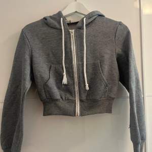 Säljer en supermysig grå croppad hoodie från Shein. Den har en dragkedja framtill och en stor luva med vita snören. Perfekt för en chill dag eller när du vill ha en avslappnad stil. Den är i storlek XS och passar perfekt till höst och vår.