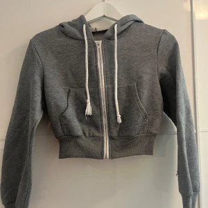 Grå croppad hoodie från Shein - Säljer en supermysig grå croppad hoodie från Shein. Den har en dragkedja framtill och en stor luva med vita snören. Perfekt för en chill dag eller när du vill ha en avslappnad stil. Den är i storlek XS och passar perfekt till höst och vår.