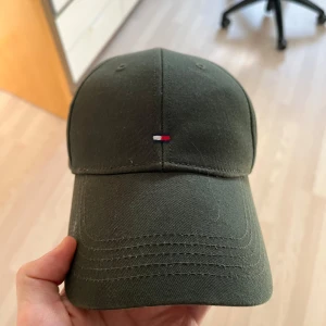 Grön keps från Tommy Hilfiger - Säljer en snygg militärgrön keps från Tommy Hilfiger. Den har en liten logga framtill och justerbar rem baktill för perfekt passform.  Simpel, och enkel. Så enkelt är det.
