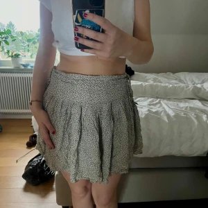 Blommig kortkjol med knytband - Säljer en superfin kortkjol med ett sött blommigt mönster. Kjolen har en bekväm passform och knytband i midjan för justering. Materialet känns lätt och luftigt.