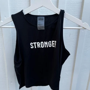 Svart ärmlös topp från Stronger - Säljer en snygg svart ärmlös topp från Stronger. Perfekt för träning eller en avslappnad dag. Tillverkad av 90% återvunnen polyester och 10% elastan, vilket gör den både bekväm och miljövänlig. Toppen har en stilren design med texten 'STRONGER' tryckt på framsidan.