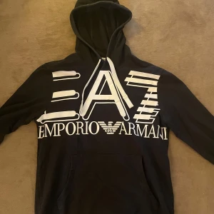 Svart hoodie från Emporio Armani - Säljer en snygg svart hoodie från Emporio Armani med stort vitt tryck på framsidan. Den har en klassisk huva och en stor ficka framtill. Det är storlek s men passar m också