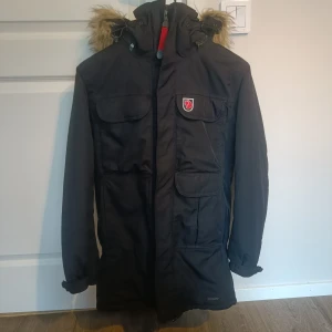 Fjällräven Kodiak vinterparka XS (mer som S) - Vinterparkas från Fjällräven. Strl XS, men stor i storlek, kan nog passa XS-M beroende på hur du vill den ska sitta - se mått på bild 3  Luva med pälskant (fuskpäls), som du kan plocka bort om du vill. Du kan även plocka bort hela huvan. Den är vind- och vattentät och ska hålla värme vid kalla förhållanden, syntetfoder gör att jackan värmer även om den skulle bli blöt (denna har dock vattenpelare på 10.000mm enl hemsida, så den ska vara rätt tålig mot väta).  Inget att anmärka på! Fynd!