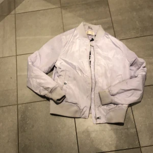 Ljuslila bomberjacka - Säljer en snygg ljuslila bomberjacka från Ü i storlek 152. Jackan har en cool text på insidan och en ficka med dragkedja på ärmen. Perfekt för vår och höst! 🧥💜