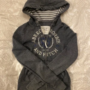 Mörkgrå hoodie från Abercrombie & Fitch - Säljer en snygg mörkgrå hoodie från Abercrombie & Fitch i storlek XS. Den har en dragkedja framtill och ett stort tryck med logga på bröstet. Perfekt för höst och vinter med sin mysiga huva och långa ärmar. Superbekväm och stilren! 