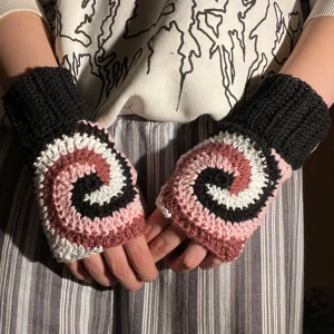 Stickade handledsvärmare med spiraldesign - Säljer ett par unika virkade handledsvärmare med en cool spiraldesign i svart, vitt och rosa. Perfekta för att hålla händerna varma under kyliga dagar. De är handgjorda och har en bekväm passform med ribbstickade muddar. Perfekt accessoar för vintern!