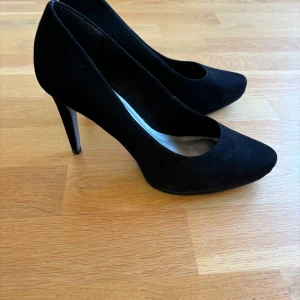 Svarta pumps från Tamaris - Snygga svarta pumps från Tamaris i mocka. De har en elegant spetsig tå och en stilren klack som passar perfekt för festliga tillfällen. Skorna är i mycket bra skick och ger en klassisk look till vilken outfit som helst.