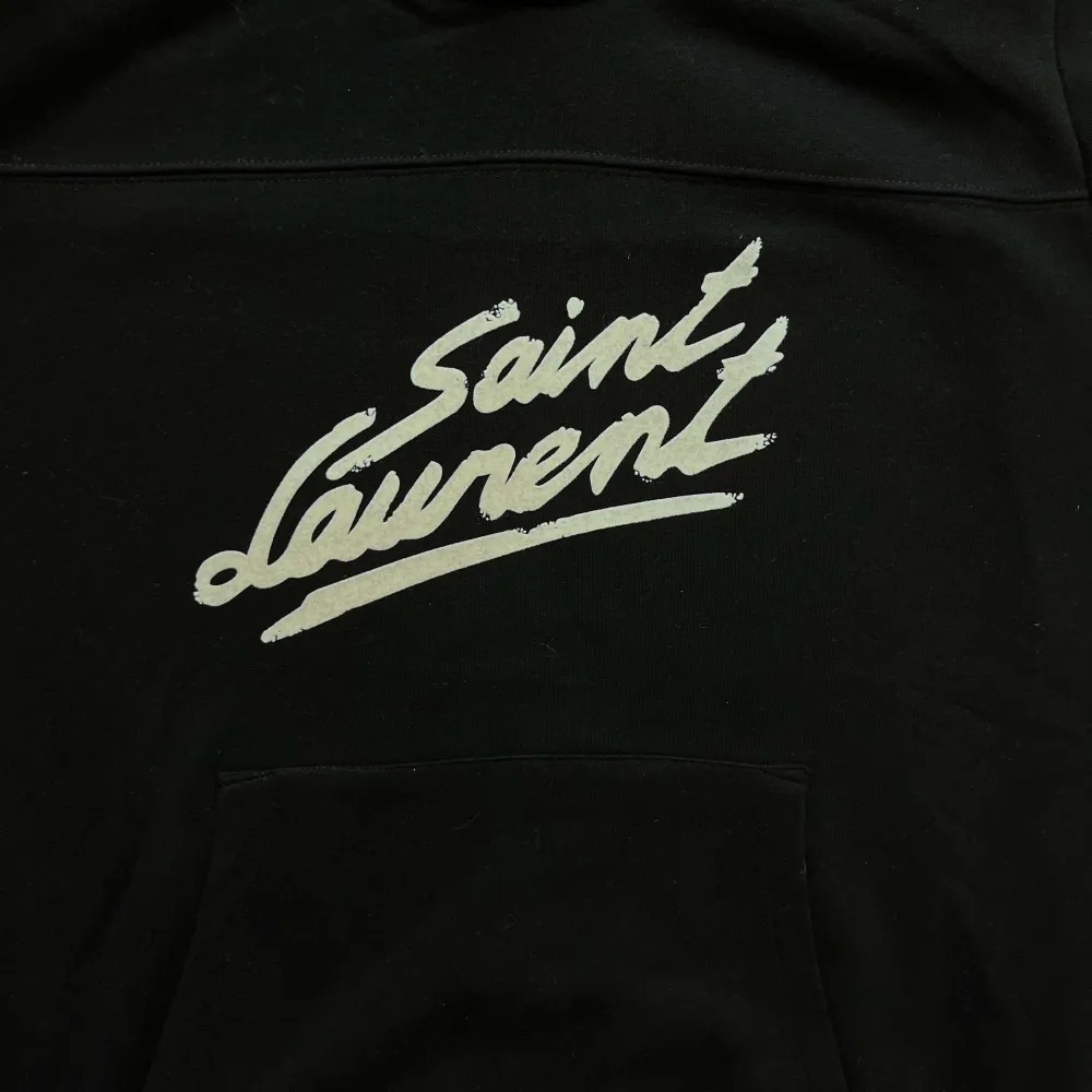 Säljer en stilren svart Saint Laurent hoodie med deras ikoniska logga i vitt på framsidan. Den har en klassisk känguruficka och justerbar huva med dragsko. Passar till allt och ger en lyxig touch till din outfit. Skriv för fler frågor.. Tröjor & Koftor.