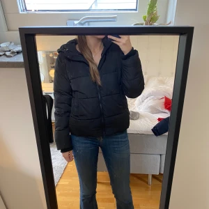 Svart pufferjacka med huva - Säljer en svart pufferjacka i nyskick. Den har en dragkedja framtill och en mysig huva. Perfekt för kalla vinterdagar och höstpromenader. Jackan är långärmad och har en normal passform. Superbekväm och stilren!