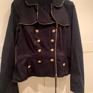 Ida Sjöstedt Svart jacka  - Snygg svart jacka från Ida Sjöstedt med guldknappar och dragkedjedetaljer. Jackan har en unik krage och dubbelknäppning framtill. Perfekt för höst och vår, med en elegant touch som passar både vardag och fest.