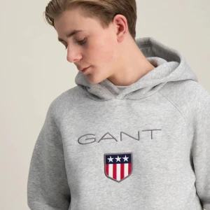 Grå hoodie från GANT - Säljer en snygg grå hoodie från GANT med deras klassiska logga och ett coolt emblem på bröstet. Perfekt för en avslappnad stil och supermjuk att ha på sig. Den har en stor ficka fram och en bekväm huva. Passar perfekt till både vardag och chill.