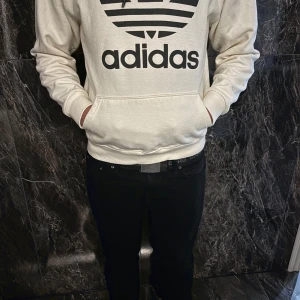 Vit hoodie från Adidas - Säljer en snygg vit hoodie från Adidas med deras klassiska logga i svart på framsidan. Den har en stor ficka framtill och en bekväm huva. Perfekt för en avslappnad stil eller träning. Passar till allt och är superbekväm! 🖤
