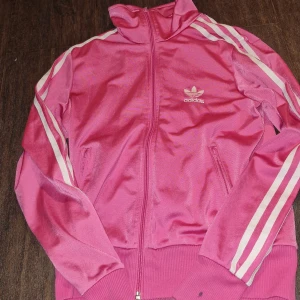 Rosa träningsjacka från Adidas - Säljer en snygg rosa träningsjacka från Adidas med klassiska vita ränder längs ärmarna. Jackan har dragkedja och en stor Adidas-logga på ryggen. Perfekt för träning eller som en cool vardagslook.