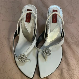 Vita klackskor  - Snygga vita sandaler från Oxogene i storlek 37. De har en elegant spetsig tå och en liten klack. Dem är dekorerade med en glittrig strassdetalj framtill som ger en lyxig touch. Perfekta för sommarens fester eller en stilfull vardag.