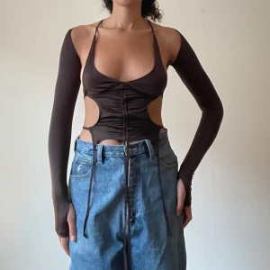 Brun snörad topp - Snygg brun topp med snörning framtill och cut-out detaljer på sidorna. Den har långa ärmar och en djup urringning, perfekt för en utekväll eller fest. Toppen är tight och ger en trendig look. Aldrig använd, tagg kvar