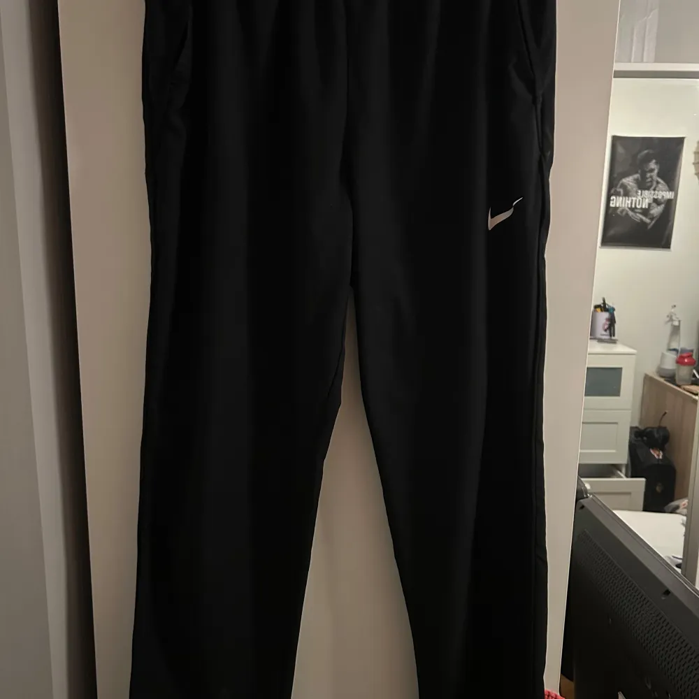 Snygga svarta träningsbyxor från Nike med en vit logga på benet. Byxorna har en normal passform och är perfekta för träning. De är tillverkade i ett syntetmaterial som ger bra rörlighet och komfort.. Farkut & Housut.