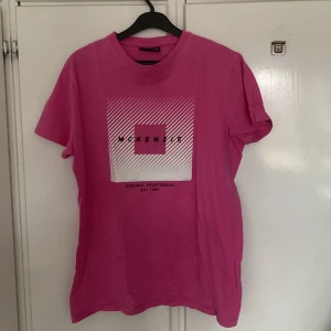Rosa t-shirt från McKenzie - Säljer en rosa t-shirt från McKenzie med tryck framtill. T-shirten har en klassisk rund hals och korta ärmar. Perfekt för en avslappnad stil. Tillverkad i ett mjukt material för extra komfort. 