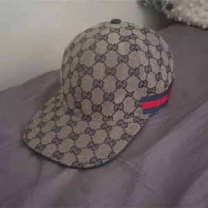 Mönstrad keps från Gucci - Snygg keps från Gucci med deras ikoniska mönster i beige och svart. Kepsen har en röd och blå randdetalj på sidan. Perfekt för att ge din outfit en lyxig touch.
