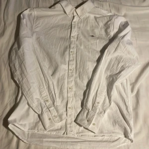 Vit skjorta från GANT - Klassisk vit skjorta från GANT med broderad logga på bröstet. Skjortan har långa ärmar och knappar framtill. Perfekt för en stilren look. Storlek 170 typ XS. Priset kan ändras om det är för högt.