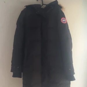 Svart dunjacka från Canada Goose - Säljer en svart dunjacka från Canada Goose med pälsdetalj på huvan. Jackan har en klassisk design med lång ärm och en broderad logotyp på ärmen. Perfekt för kalla vinterdagar. 🖤Har även tillbehör (påse)