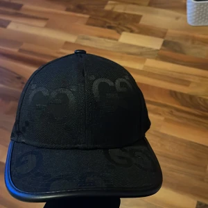 Svart keps från Gucci - Snygg svart keps från Gucci med diskret mönster av märkets logotyp. Kepsen har en klassisk design med böjd skärm och justerbar passform. Perfekt för att ge en stilren touch till din outfit.
