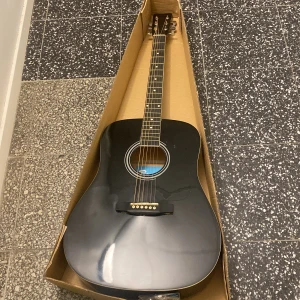 Svart akustisk gitarr från Rusta - Säljer en stilren svart akustisk gitarr från Rusta. Perfekt för nybörjare och inkluderar tillbehör som stämapparat och plektrum. Gitarren har en klassisk design med sex strängar och levereras i originalförpackning. På baksidan av gitarren finns små skrapor. Jag har inte använt så mycket, har ingen användning till den det är  just därför jag säljer den. Ett bra val för den som vill börja spela eller till den som redan spelar gittar!🎸