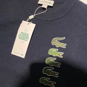 Mörkblå tröja från Lacoste med krokodiler - Snygg mörkblå tröja från Lacoste med flera ikoniska gröna krokodiler broderade på framsidan. Tröjan har en rund halsringning och är perfekt för en stilren look.